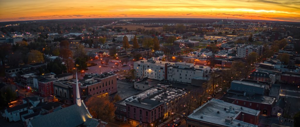 View of a small town at sunset from an elevated perspective, showcasing illuminated streets, buildings, and a church steeple in the foreground, with a glowing orange and yellow sky in the background.