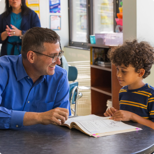 Governor Matt Meyer in a blue shirt smiling and engaging with a young student who is reading a book at a classroom table.