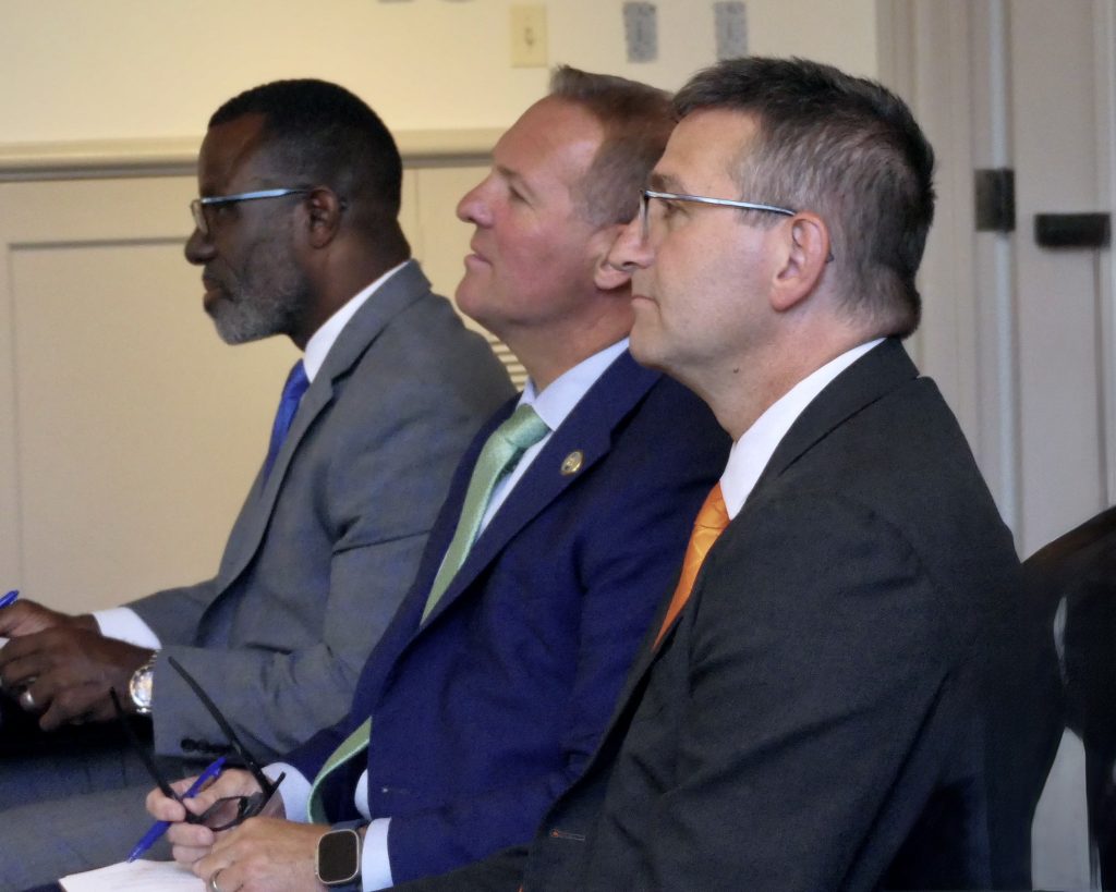 Alt text: Three men in suits sit side by side at a meeting or panel, shown in profile from the right. Each is wearing a tie and glasses, holding a pen and notepad, and looking attentively toward something off camera. The setting appears to be an indoor conference or official meeting room with neutral walls and doors in the background.