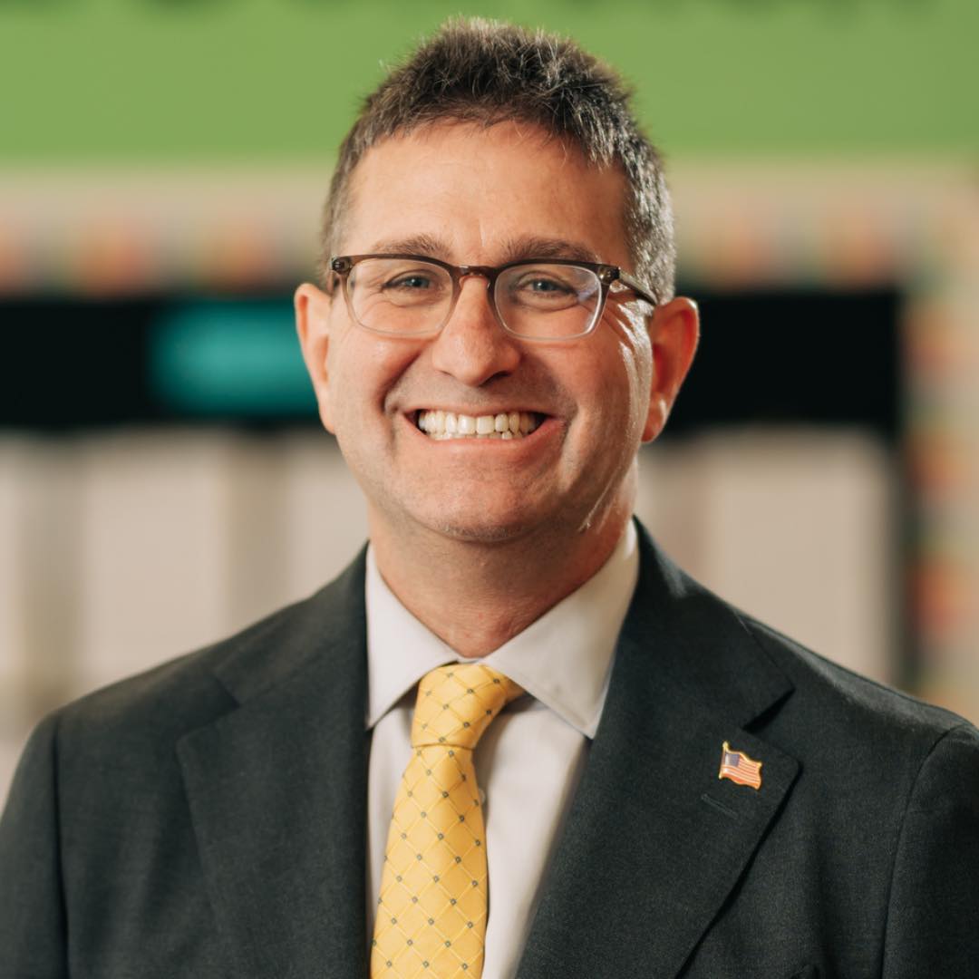 Governor Meyers wears glasses, a dark suit, white shirt, and a yellow patterned tie with an American flag pin on his lapel, standing indoors against a softly blurred background.