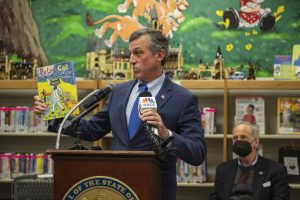Governor Carney stands behind a podium holding a book while inside a library.