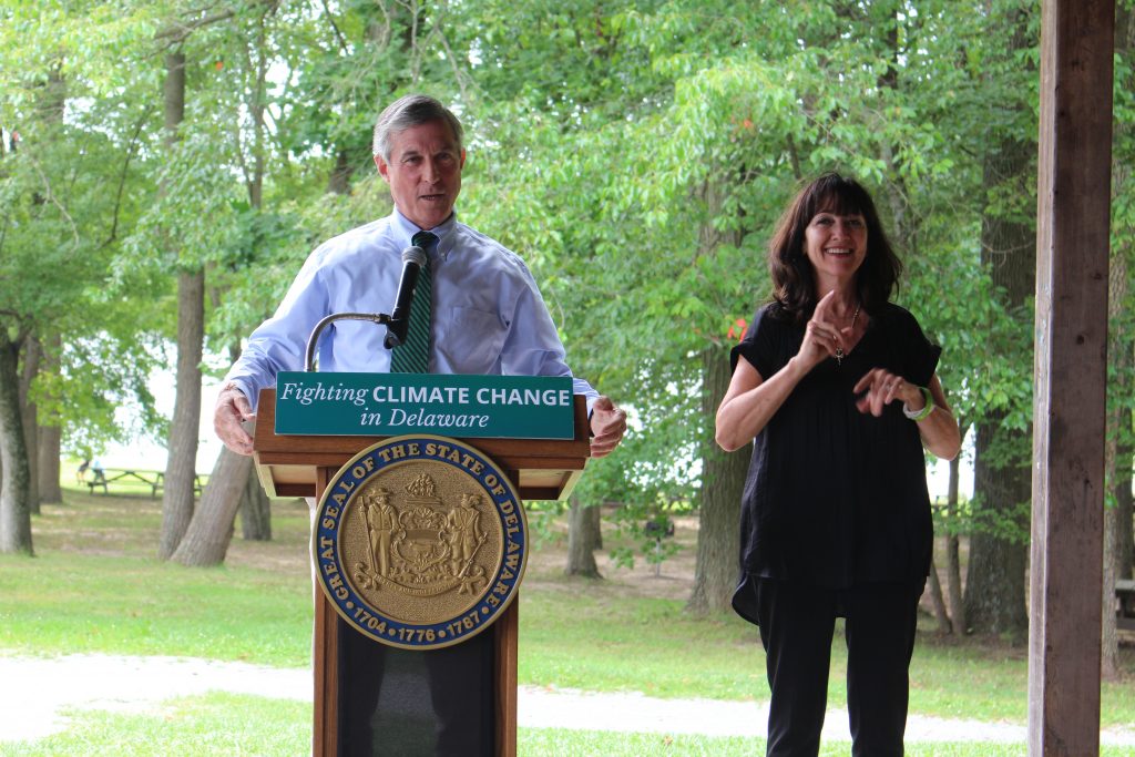 Governor Carney delivers remarks from behind a podium, while an ASL intrepreter stands off to the side. On the podium is the state seal and a sign that reads Fighting Climate Change in Delaware.
